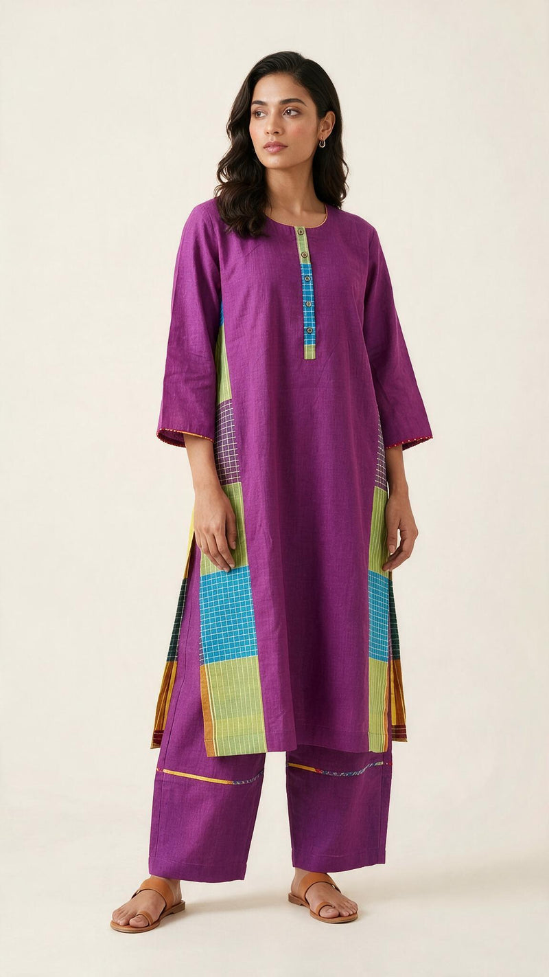 A woman modeling a purple three-piece kurta set with colorful patchwork details in blue, green, and yellow, photographed against a plain light beige studio background.