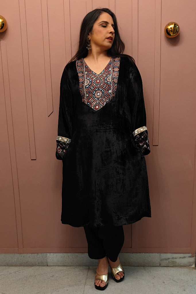 Woman wearing a black traditional outfit with intricate patterns in front of a pink wall.